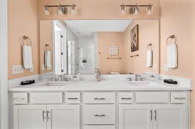 Bright residential bathroom with white double-sink vanity and cabinets, white quartz countertop, large wall mirror, brushed-nickel faucets and lights, peach-colored walls, towel rings and simple decor.