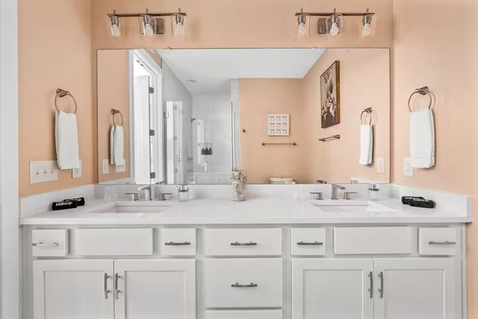 Bright residential bathroom with white double-sink vanity and cabinets, white quartz countertop, large wall mirror, brushed-nickel faucets and lights, peach-colored walls, towel rings and simple decor.
