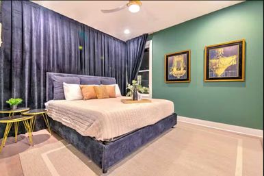Contemporary bedroom interior with navy velvet platform bed, beige quilt and pillows, floor-to-ceiling dark curtains, green accent wall with two framed gold chandelier prints, gold side tables and neutral rug.