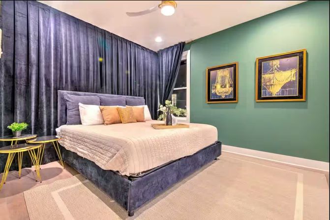Contemporary bedroom interior with navy velvet platform bed, beige quilt and pillows, floor-to-ceiling dark curtains, green accent wall with two framed gold chandelier prints, gold side tables and neutral rug.