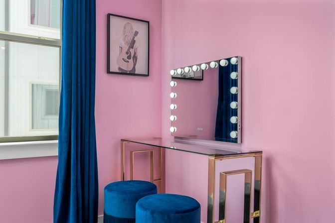 Pink interior makeup vanity nook with glass and gold-accented table, illuminated Hollywood mirror, deep blue velvet stools and matching curtain, framed guitar art by the window