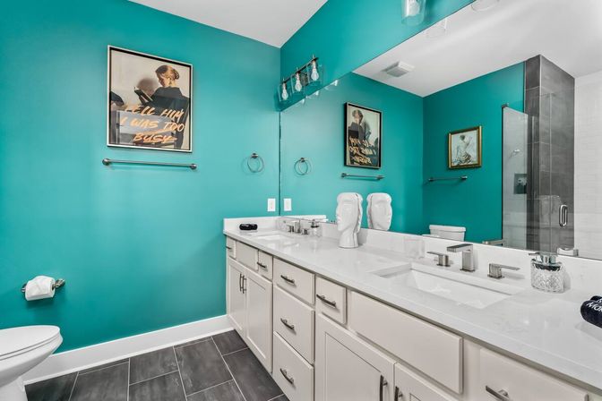 Contemporary turquoise home bathroom with white double vanity and quartz countertop, chrome faucets, large mirror, glass-enclosed shower, framed wall art and charcoal floor tiles.
