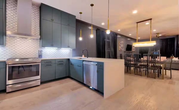 Modern open-concept kitchen and dining area with gray cabinets, white waterfall island, stainless steel oven and dishwasher, patterned backsplash, pendant lighting and a dining table with black chairs on light hardwood floors.
