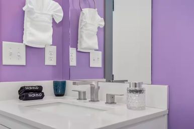 Cheerful purple bathroom vanity with white sink and modern chrome faucet, stacked 'Makeup' towels, blue tumbler and textured glass jar