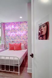 Bright pink-themed bedroom interior with a white metal bed frame, coral throw pillows, pastel geometric wallpaper and a neon pink script sign that reads And by the way I'm going out tonight.