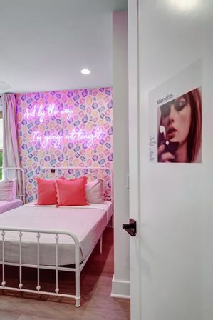 Bright pink-themed bedroom interior with a white metal bed frame, coral throw pillows, pastel geometric wallpaper and a neon pink script sign that reads And by the way I'm going out tonight.