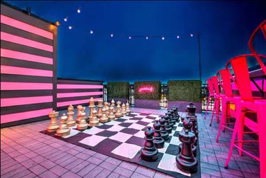 Urban rooftop bar at night featuring an oversized chessboard with giant gold and black pieces, pink neon-striped wall, string lights, and bright pink bar stools.