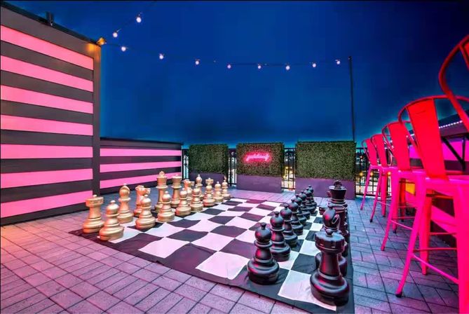 Urban rooftop bar at night featuring an oversized chessboard with giant gold and black pieces, pink neon-striped wall, string lights, and bright pink bar stools.