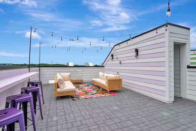 Sunny rooftop patio with string lights, pastel-striped siding, wicker sofas on a floral rug and purple bar stools