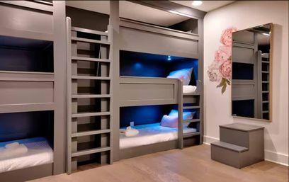 Gray built-in bunk beds with ladders and blue LED reading lights in a modern kids’ bedroom, white bedding, floral wall decal, full-length mirror and small step stool on hardwood floor.