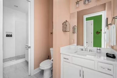 Peach-walled residential bathroom with white vanity and sink, glass walk-in shower, toilet, wire wall baskets and mirror reflecting a green hallway.