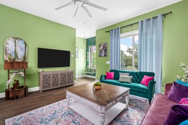 Sunlit modern living room interior with lime-green walls, teal velvet sofa and pink pillows, mauve loveseat, wooden coffee table on a floral rug, wall-mounted TV and large window with blue curtains overlooking trees.