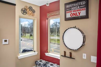 Music-themed studio corner with two windows overlooking a street and park, red accent wall with lit 'SHOWTIME' marquee, mounted drum and 'Restricted Area: Band Members Only' sign, film-reel wall art and patterned bench.