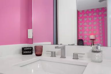 Playful pink bathroom vanity with white quartz sink, modern chrome faucet, makeup towels and a purple tumbler on the counter, mirror reflecting pink patterned wallpaper and a lamp.