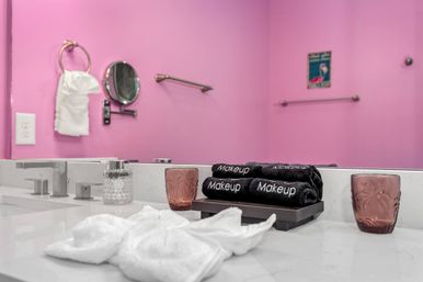 Playful pink bathroom vanity with marble countertop, dual faucets, stacked black towels labeled Makeup on a tray, rose glass tumblers, white washcloths, magnifying mirror and towel ring.