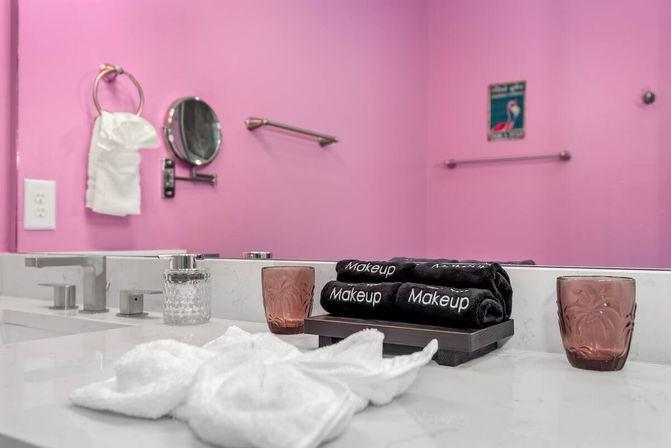 Playful pink bathroom vanity with marble countertop, dual faucets, stacked black towels labeled Makeup on a tray, rose glass tumblers, white washcloths, magnifying mirror and towel ring.