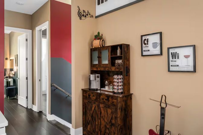 Modern home hallway coffee station with rustic dark-wood cabinet and single-serve coffee maker, beige walls with red and gray accent panels, framed coffee and wine prints, a music-note wall decal, dark hardwood floors and a guitar on a stand.