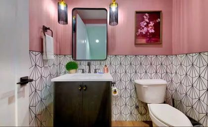Compact modern pink powder room with geometric black-and-white tile, dark vanity, rectangular mirror, pendant lights, white sink and toilet, potted plant and floral wall art.