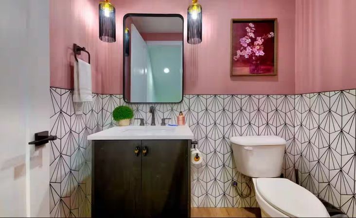Compact modern pink powder room with geometric black-and-white tile, dark vanity, rectangular mirror, pendant lights, white sink and toilet, potted plant and floral wall art.