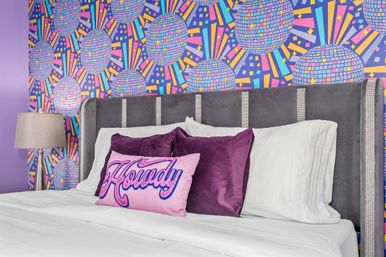 Playful modern bedroom interior with gray upholstered headboard, crisp white bedding, purple velvet pillows and pink 'Howdy' accent pillow against colorful disco-ball patterned wallpaper with a bedside lamp.