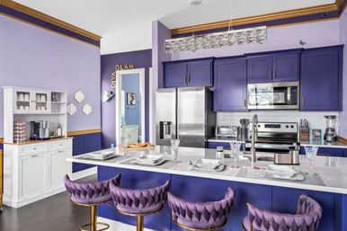 Glam modern purple kitchen with lavender walls and deep plum cabinets, white quartz island set for dining, tufted velvet purple bar stools with gold bases, crystal pendant light, and stainless steel refrigerator, oven and microwave.