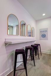 Pastel pink interior featuring a white floating counter, four gold-framed arched mirrors and four glossy purple metal bar stools on a light wood floor — modern vanity station.