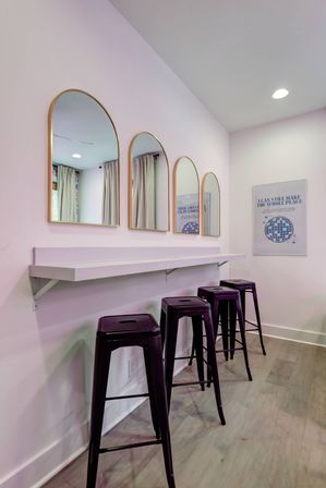 Pastel pink interior featuring a white floating counter, four gold-framed arched mirrors and four glossy purple metal bar stools on a light wood floor — modern vanity station.