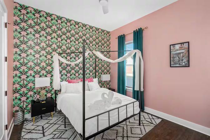Bright boutique-style bedroom with black metal canopy bed draped in white fabric, white linens and towel swans, green-and-pink art-deco wallpaper accent wall, teal curtains, pink painted walls, black nightstand and geometric rug.
