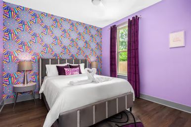 Colorful purple guest bedroom with geometric patterned accent wall, gray upholstered bed dressed in white linens with towel swans, purple pillows and curtains, bedside lamps, window showing green yard, and dark wood floor.