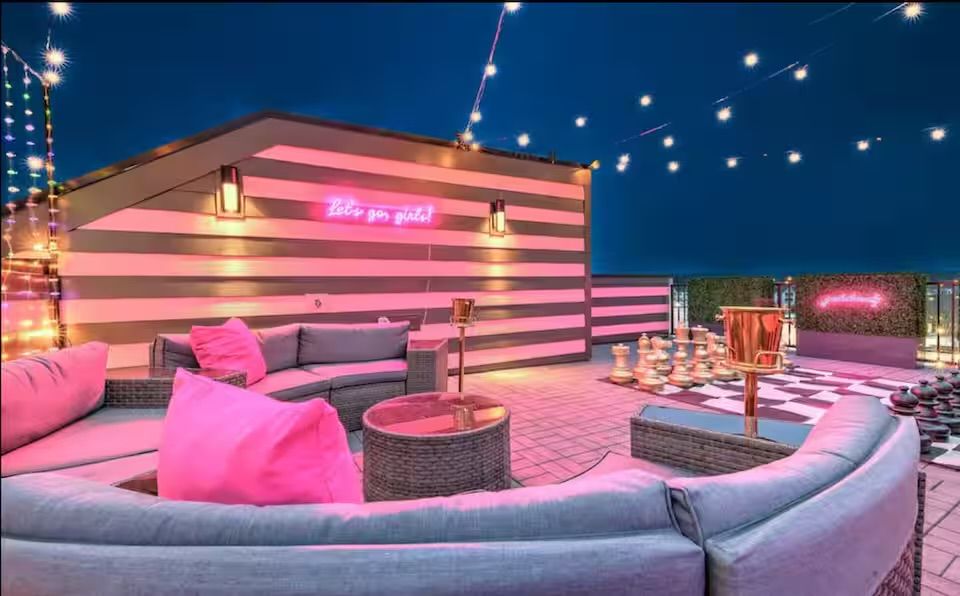 Urban rooftop lounge at dusk with curved wicker sectional and bright pink cushions, striped accent wall and pink neon sign, string lights and oversized outdoor chess set.