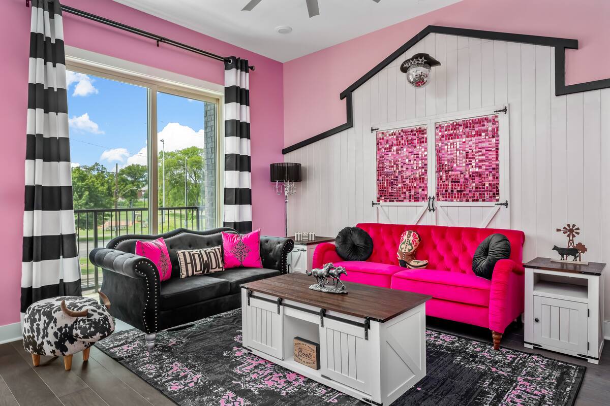 Vibrant pink living room with hot-pink tufted sofa, black velvet Chesterfield couch, black-and-white striped curtains, farmhouse coffee table and pink mosaic barn-door wall art