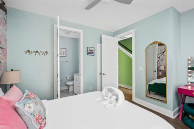 Bright pastel bedroom with light-blue walls, white bed topped with pink and floral pillows and a towel swan, open doors revealing an en-suite bathroom and a bright green stairwell, gold-framed full-length mirror, pink vanity and ceiling fan — coastal-inspired decor.