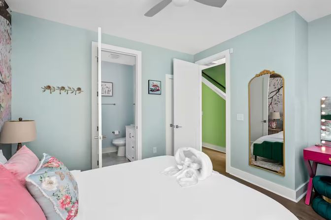 Bright pastel bedroom with light-blue walls, white bed topped with pink and floral pillows and a towel swan, open doors revealing an en-suite bathroom and a bright green stairwell, gold-framed full-length mirror, pink vanity and ceiling fan — coastal-inspired decor.