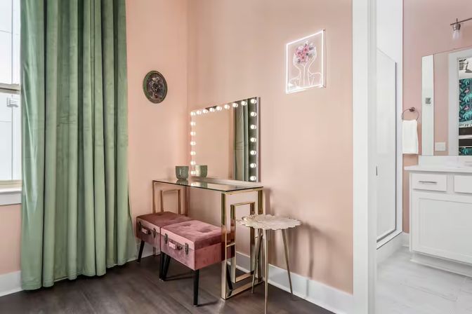 Glam makeup vanity corner with lighted Hollywood mirror, glass-and-gold desk, pink velvet stools, small gold side table, blush-pink walls and green floor-length curtains with bathroom doorway visible.
