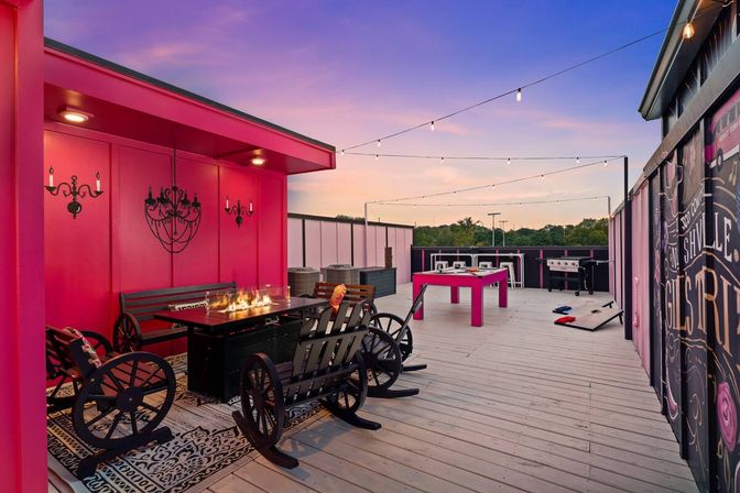 Rooftop deck at sunset with vibrant pink walls and string lights, wagon-wheel rocking chairs around a fire-pit table, pink ping-pong table, cornhole boards and grills on a wood deck.