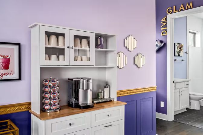 Cozy lavender and purple salon beverage station with white cabinets, wood countertop, espresso machine and K‑Cup carousel, decorative gold trim and small mirrors, doorway to a restroom