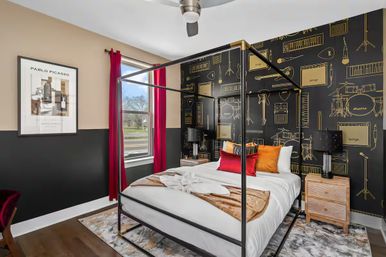 Stylish modern bedroom with black metal four-poster bed, white linens and red and orange accent pillows, music-themed black-and-gold wallpaper with instrument illustrations, red curtains at a sunny window, wooden nightstands and lamps on dark hardwood floors.