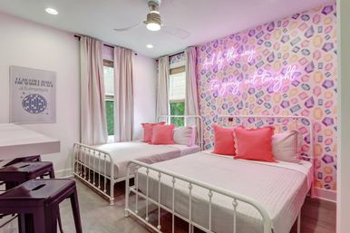 Cheerful modern twin bedroom with two white metal beds, pink throw pillows, pastel geometric accent wall topped by a pink neon script sign, tall windows with pale curtains, ceiling fan, and light wood floors.