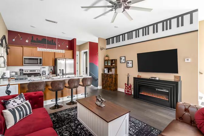 Nashville-inspired open-plan living room and kitchen with red sofa, white island and four brown bar stools, stainless steel appliances, wall-mounted TV above electric fireplace, decorative guitars and piano-key wall art, large ceiling fan