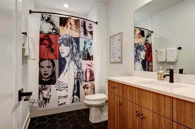 Contemporary residential bathroom with wood vanity and white countertop, black hexagon floor tile, large mirror, toilet, and a colorful pop‑star portrait collage shower curtain for a playful contrast.