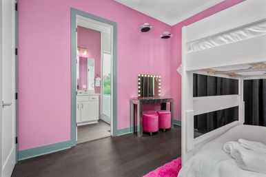 Modern pink kids' bedroom with white bunk bed, lighted vanity mirror and two velvet pink stools, dark hardwood floor and doorway to a bathroom