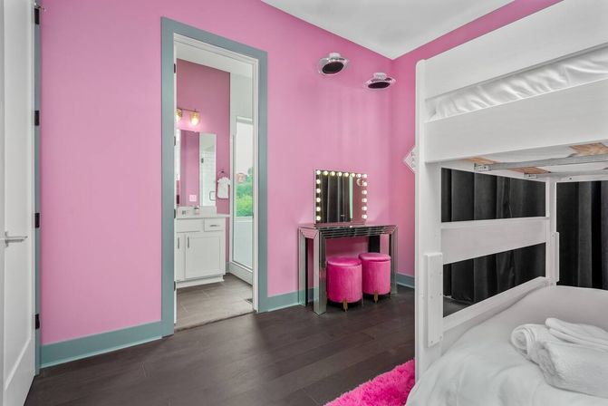 Modern pink kids' bedroom with white bunk bed, lighted vanity mirror and two velvet pink stools, dark hardwood floor and doorway to a bathroom