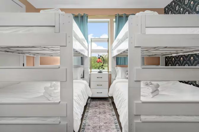 Bright family bunk room with two white twin-over-twin bunk beds facing each other, crisp white linens and folded towels, narrow patterned runner leading to a dresser and lamp under a window with teal curtains.