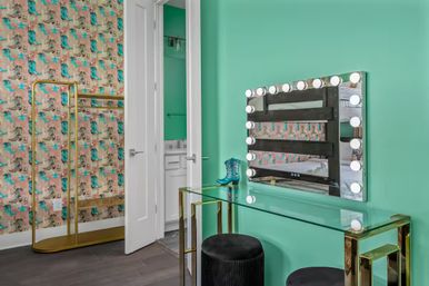 Mint-green dressing room with glass-and-gold vanity, Hollywood-style lighted mirror, black velvet stools, patterned teal-and-pink wallpaper, gold clothing rack and an open door to a white bathroom.