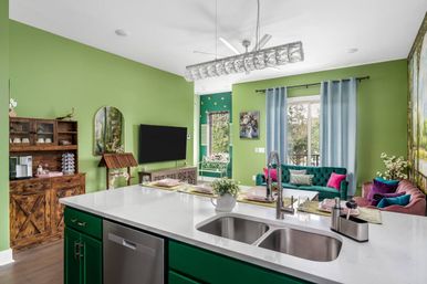 Sunlit open-concept kitchen and living room with bright green walls, white quartz island featuring a double sink and stainless dishwasher, dark green base cabinets, rustic wooden hutch, wall-mounted TV, teal and magenta velvet sofas, light blue curtains and a large window