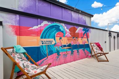 Beach-themed mural of pink flamingos, a teal ocean wave and colorful sunset painted on a building wall above a white wooden deck with tropical-patterned lounge chairs and string lights.
