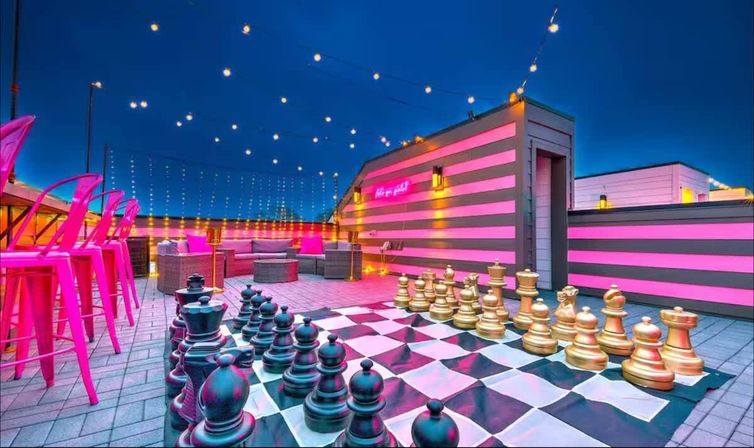 Urban rooftop lounge at dusk with oversized black and gold chess set on a checkerboard, pink neon-striped walls, string lights and hot-pink bar stools.