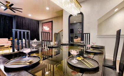 Glamorous contemporary dining room with a glossy black table set with black chargers, gold flatware and pink dessert coupes, tall black chairs, crystal chandelier, and open-plan living area with velvet sofas and dark drapes.
