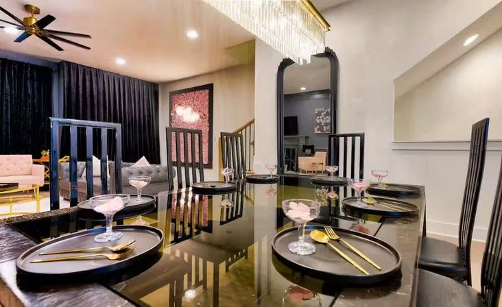Glamorous contemporary dining room with a glossy black table set with black chargers, gold flatware and pink dessert coupes, tall black chairs, crystal chandelier, and open-plan living area with velvet sofas and dark drapes.