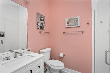 Cheerful modern pink bathroom with white vanity and sink, chrome fixtures, glass shower, toilet, towel bars and retro wall art.